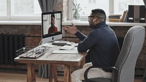 Middle eastern man in wireless earphones sitting at desk in loft office and speaking with African American woman via video call on computer while having online business meeting or lesson