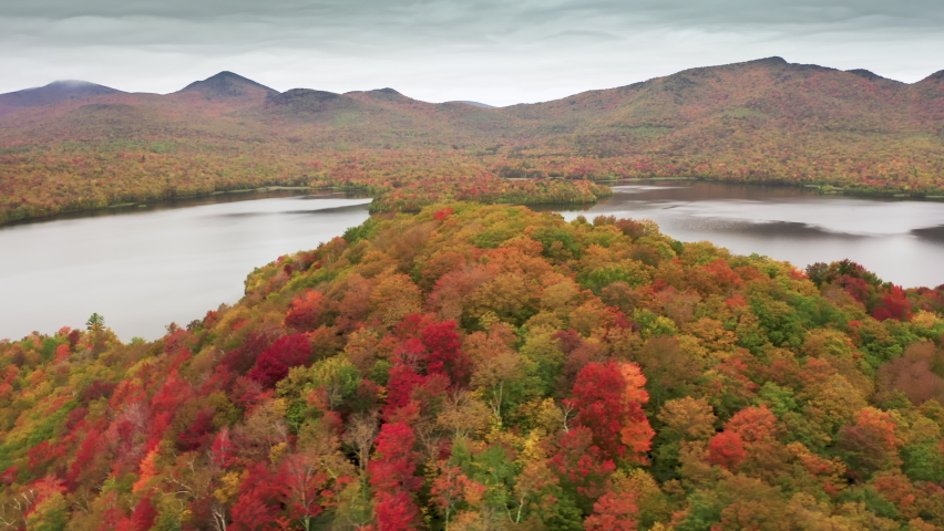 Bird eye view on colorful forrest hill with mixed trees turning leaves into yellow, orange and red vibrant colors. Scenic wilderness nature lake on cloudy moody rainy day at Vermont, New England, USA