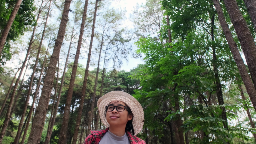 Hipster woman in straw hat and red plaid shirt is walking in a pine alley enjoying nature. Happy Asian female traveler sightseeing in pine tree garden.