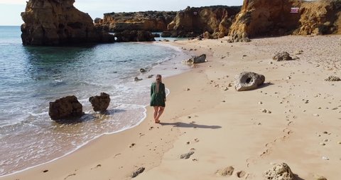 Woman in beautiful waving dress walking at the empty Algarve beach. Drone taking off and going over and up with the camera. Walking on the golden beach with crystal clear water and rocky coast