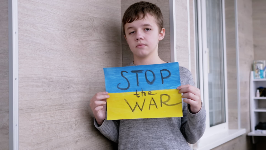 Child Holds in Hands a Poster with a Flag of Ukraine, and Message Stop the War. Sad boy calling protests against military conflict between Russia and Ukraine in a drawing with a blue and yellow flag. Royalty-Free Stock Footage #1088847283
