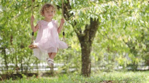 Spring and happy summer time. Joyful smiling little girl swinging on the swing, child with blue eyes and curly blond hair plays in the green garden at home, concept of healthy growth