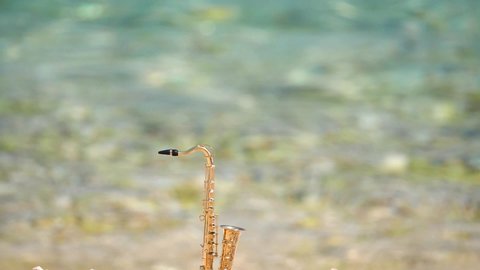 Mini model of real alt tenor saxophone stands white small pebbles on seashore, against background of blue turquoise water. Music screensaver for romance. Slow motion video. Copy space for your text.