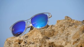 Mirrored blue and turquoise sunglasses lie on beige sea stones, on sea, on seashore, against backdrop of blue sky. Slow motion video. The concept of summer vacation, rest and relaxation.