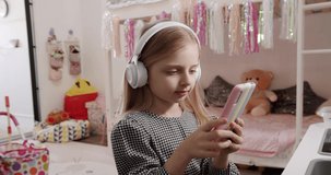 The 7-year-old girl is playing in the children's room with white headphones and using colored smartphone. enjoying cool video or photo content in social network, playing online games.