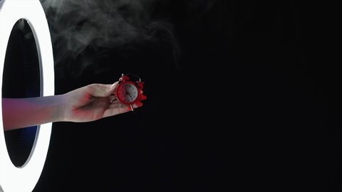 Final sale. Last minute offer. Hurry up. Time reminder. Female hand shaking red alarm clock in smoke cloud LED light ring isolated on dark night black copy space background.