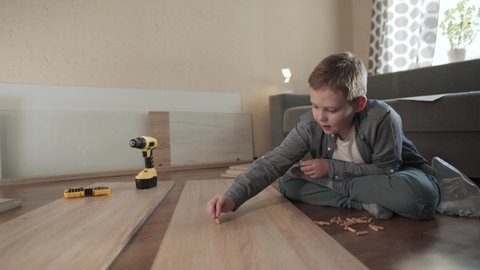Joyful caucasian boy sits on floor and inserts wooden dowels into chipboard panel while self-assembling furniture. Warm room with laid out pieces of furniture and tools. 