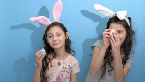 4k. Happy easter. Cute girls sisters in rabbit bunny ears on head holding painted easter eggs on blue background, going to celebrate Easter. Spring holiday concept