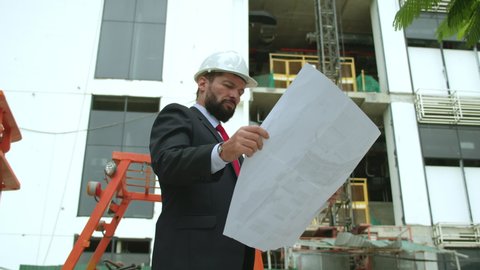 Portrait of a successful handsome engineer of bearded, senior architect, builder, businessman in white helmet, in suit, holding a project in his hand,background of a skyscraper and a construction site