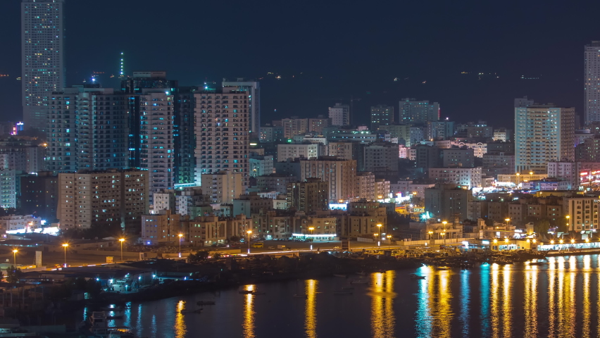 Cityscape of Ajman aerial view from rooftop at night timelapse with reflection of water in lake. Illuminated towers on a waterfront in the United Arab Emirates.