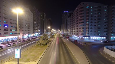Cityscape of Ajman with traffic on road between many towers Aerial view from a bridge with lights in illuminated buildings at night timelapse. United Arab Emirates.