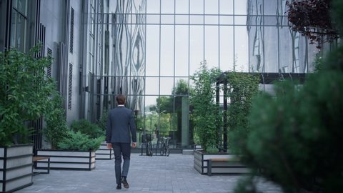 Confident businessman walking downtown area rear view. Modern glass office building facade with neat landscaped garden. Successful man director going workplace in suit. European financial institution.