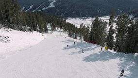 Aerial view of a ski resort with people snowboarding and skiing from a hill. Video footage. Flight over ski or snowboard track on white snow surrounded by thick forest