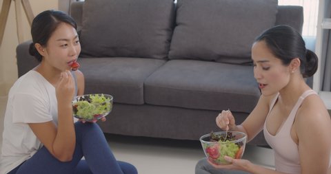 Two beautiful Asian woman eats  salad  from a bowl with sitting on the floor in the living room.Eat nutritious food.Yoga woman.Slow motion.Healthy lifestyle  concept.