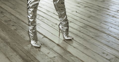 Feet of women dressed in high heeled boots of silver color move along the wooden floor, backlight