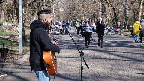 Russia, Essentuki, March 10, 2022. A young street musician is playing guitar in a city park on a sunny spring day, people are walking in the park. The concept of urban lifestyle, street musicians