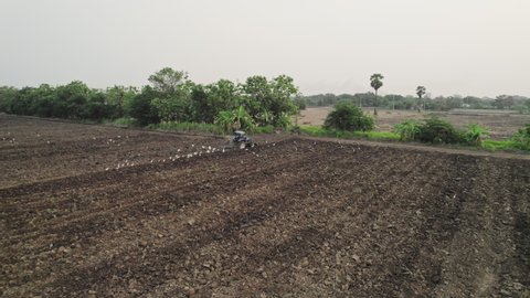Tractor machine plowing ground on farm field of dust trails behind preparing soil for planting new crop, agriculture concept, top view, drone shooting.