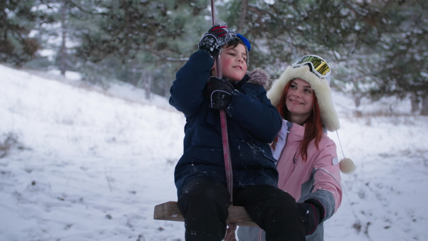 family weekend in snowy weather, a charming woman together with her son has fun riding a makeshift swing in a winter forest among trees