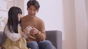 Happy Asian father teaching daughter to play games with joy stick on sofa in relax day.