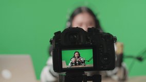 Close Up Of A Camera Monitor Recording A Smiling Asian Woman Gamer With Headphone And Computer Looking At Camera While Playing Mobile Phone Game On The Green Screen 
