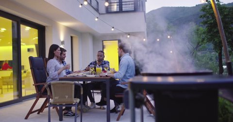 A group of friends enjoy while having dinner together in front of a luxury villa. A group of friends enjoy the barbecue and have fun in the late hours