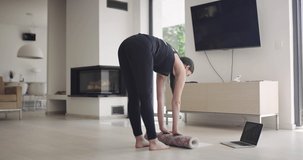 woman in sportswear doing stretching exercises while watching yoga training class on computer laptop online. Healthy girl exercising in living room with sofa couch in the background..