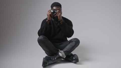 African American man sitting legs-crossed on the floor and taking a photo with camera full shot studio shot grey background copy space. High quality 4k footage