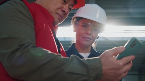 At sunlight portrait two factory technicians ith helmet use mobile phone standing in factory. Woman and man. Engineers, team