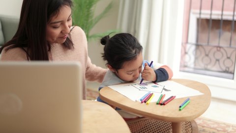 Asian mother taking care of her child painting while using laptop computer at home - Family lifestyle