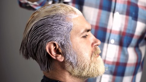 Dyed hair for a bearded hipster guy. Hairdresser applying dye to man hair. Hair coloring in gray color process. Process of a guy having beard and moustache dyed, close up.