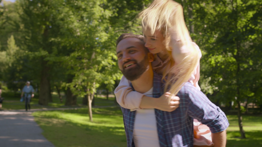 Happy summer date. Carefree middle aged couple in love playing together, man piggybacking laughing woman in green park, tracking shot, slow motion