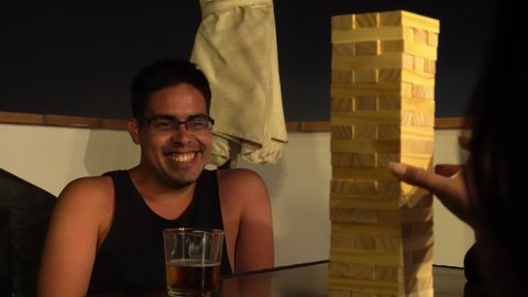 close-up of a man watching in disbelief and nervous and smiling a board game of wooden blocks pulling out with his hand in a meeting on the terrace with friends with beer at night in 4k