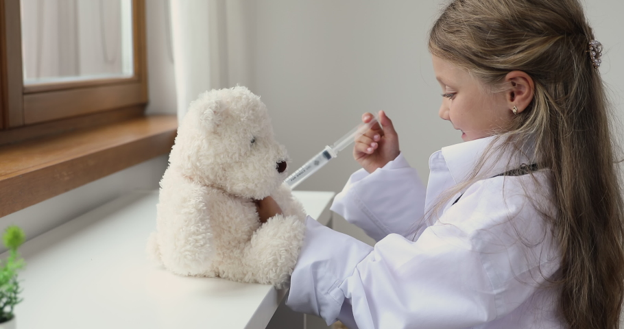Little girl wear white coat play pediatrician, pretend be doctor or nurse treats fluffy bear holds toy syringe, make injection or vaccine, have fun at home. Healthcare, medicine, vet clinic ad concept Royalty-Free Stock Footage #1088885217