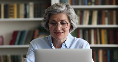 Close up smiling attractive older 50s businesslady in glasses working on laptop sit at desk in modern office look concentrated and motivated. Busy workday, workflow using modern tech, blogging concept