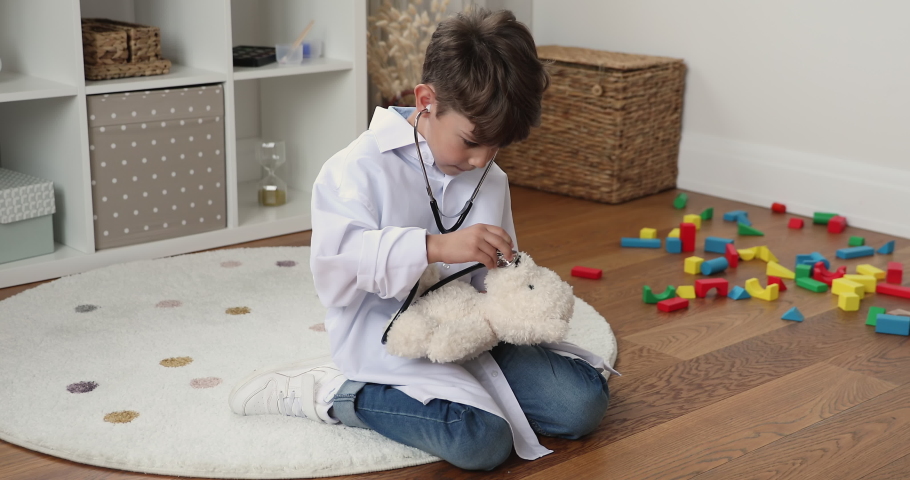 Little 6s boy wear white coat play veterinarian use stethoscope listen plush bear heartbeat, make medical checkup, imagines his doctor, having fun sits on floor in modern nursery. Health-care concept Royalty-Free Stock Footage #1088885379