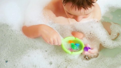 A cute baby boy fishes colorful toys out of the bath and puts them in a green small ladle close-up