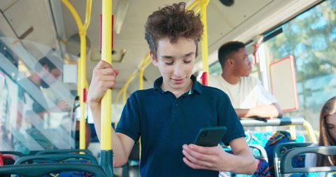 Young school-age boy rides public transportation bus to elementary school holding phone in hand, looks at smartphone while holding onto handrail, waits for bus to stop so he can get off