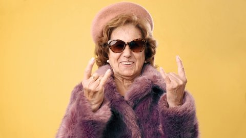 Stylish senior woman making a rock n roll gesture with her fingers over an yellow background