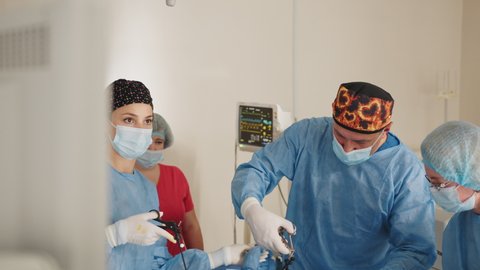 Portrait of female medical worker in protective mask and sterile gloves holding laparoscopic grasper with trocar while standing near table with surgical tools. Surgeon and his assistant on blurred.