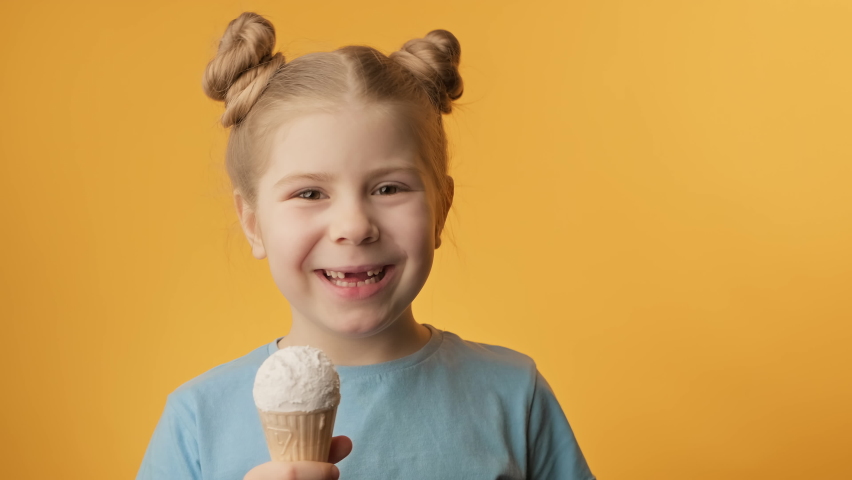 Toothless girl eating ice cream cone on yellow background. Cute toothless little girl enjoys delicious vanilla ice cream. Close-up in 4K, UHD | Shutterstock HD Video #1088887865