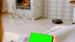Robot vacuum cleaner removes dust on laminate floor near modern fireplace. Woman rests on couch watching videos in mobile phone close view