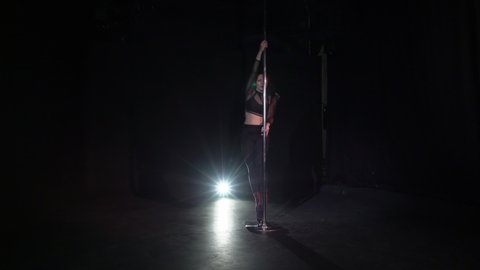 Pole dance. Young beautiful caucasian brunette woman dancer in studio at black background. Girl dancing striptease. Sexy female in shoes on high heels strips, pants and crop top rotates and twirls.