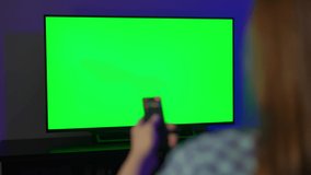A young woman sits on an armchair and watches TV. Turns on, switches channels with the remote control in the evening or at home at night. Green screen isolated chroma key.