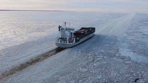 A large ship with cargo containers goes along the sea fairway in the middle of the ice in winter at sunset, aerial view from behind. Russia, St. Petersburg, January 2022