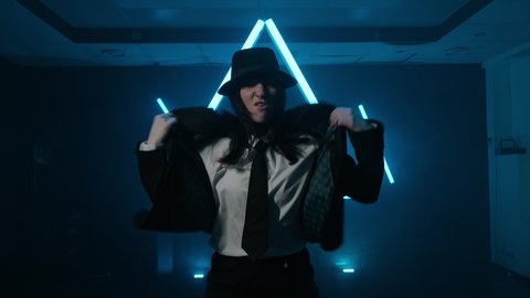 Young woman dancing hip hop in smoky studio. Professional dancer in suit and hat dancing hip hop in dark studio. Cyberpunk. Youth culture. Talented female dancer. Triangle shape at background