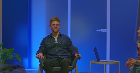 Young blogger on a talk show, speaks emotionally and gesticulates. Red-haired man in glasses sits in a chair, modern business center. 4k, ProRes