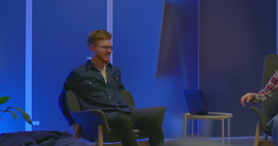 Red-haired man at a meeting of bloggers in a modern coworking center, talk show. Young man with glasses is sitting in a chair, talking with colleagues and laughing. 4k, ProRes