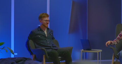 Red-haired man at a meeting of bloggers in a modern coworking center, talk show. Young man with glasses is sitting in a chair, talking with colleagues and laughing. 4k, ProRes