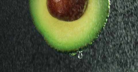 Cinematic macro shot of half raw fresh organic tropical green avocado fruit with falling water drops isolated on black background.