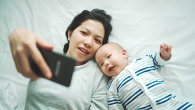 Mom and her son making video call with father and family, Happy Asian Young mother and her little baby boy lying on bed smiling face waving hand talking to smartphone